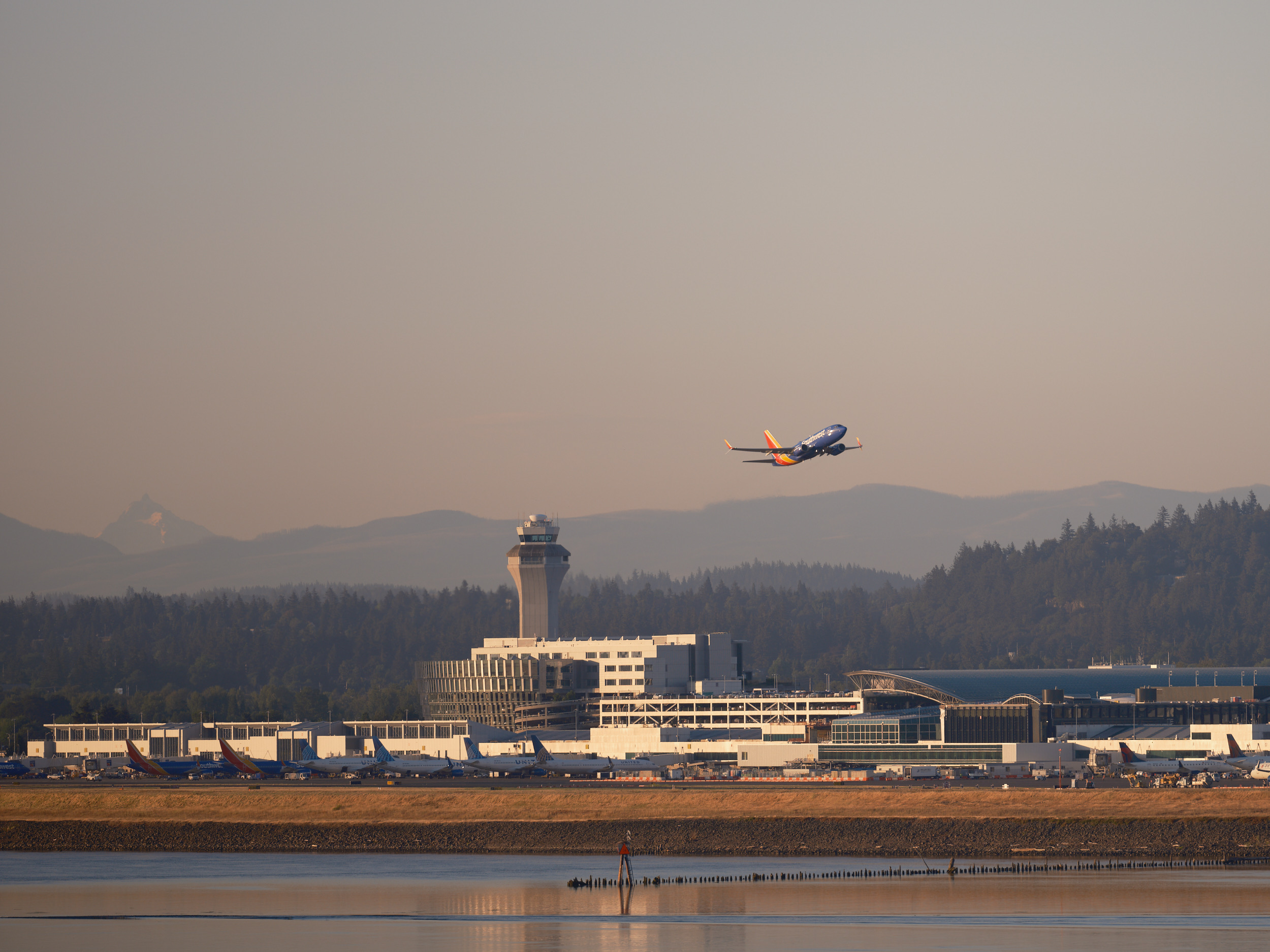 A Southwest Airlines aircraft takes off against a dramatic sunset sky above the Portland International Airport terminal and control tower, with forested hills and mountains visible in the background and calm water in the foreground.