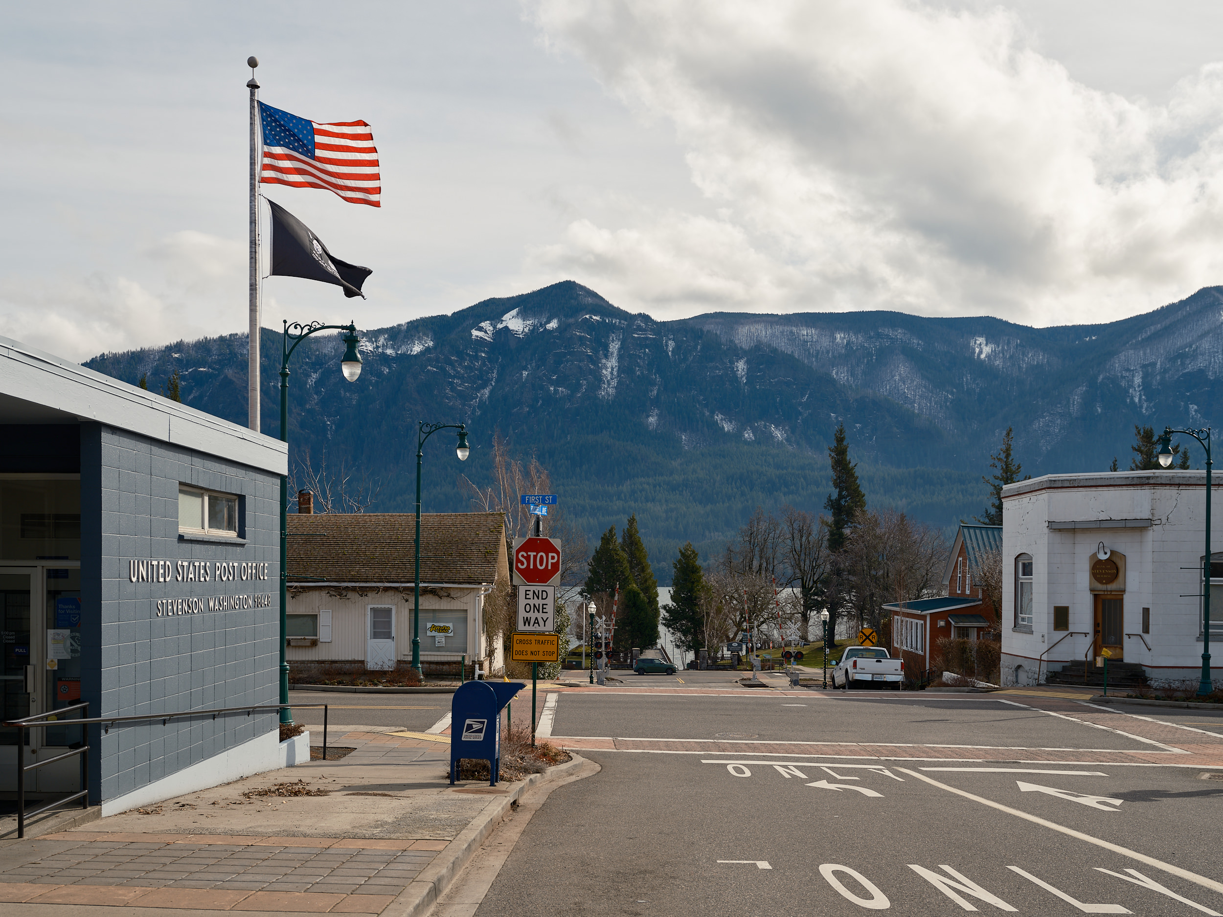 The United States Post Office in Stevenson, Washington, a single-story blue brick building with signage reading "Stevenson Washington 98648." An American flag and a black POW/MIA flag fly from a tall flagpole. A blue USPS mailbox sits on the sidewalk near a stop sign and "End One Way" sign at an intersection. In the background, a quiet small-town main street leads toward the Columbia River Gorge, with densely forested mountains dusted with snow rising dramatically under a partly cloudy sky.