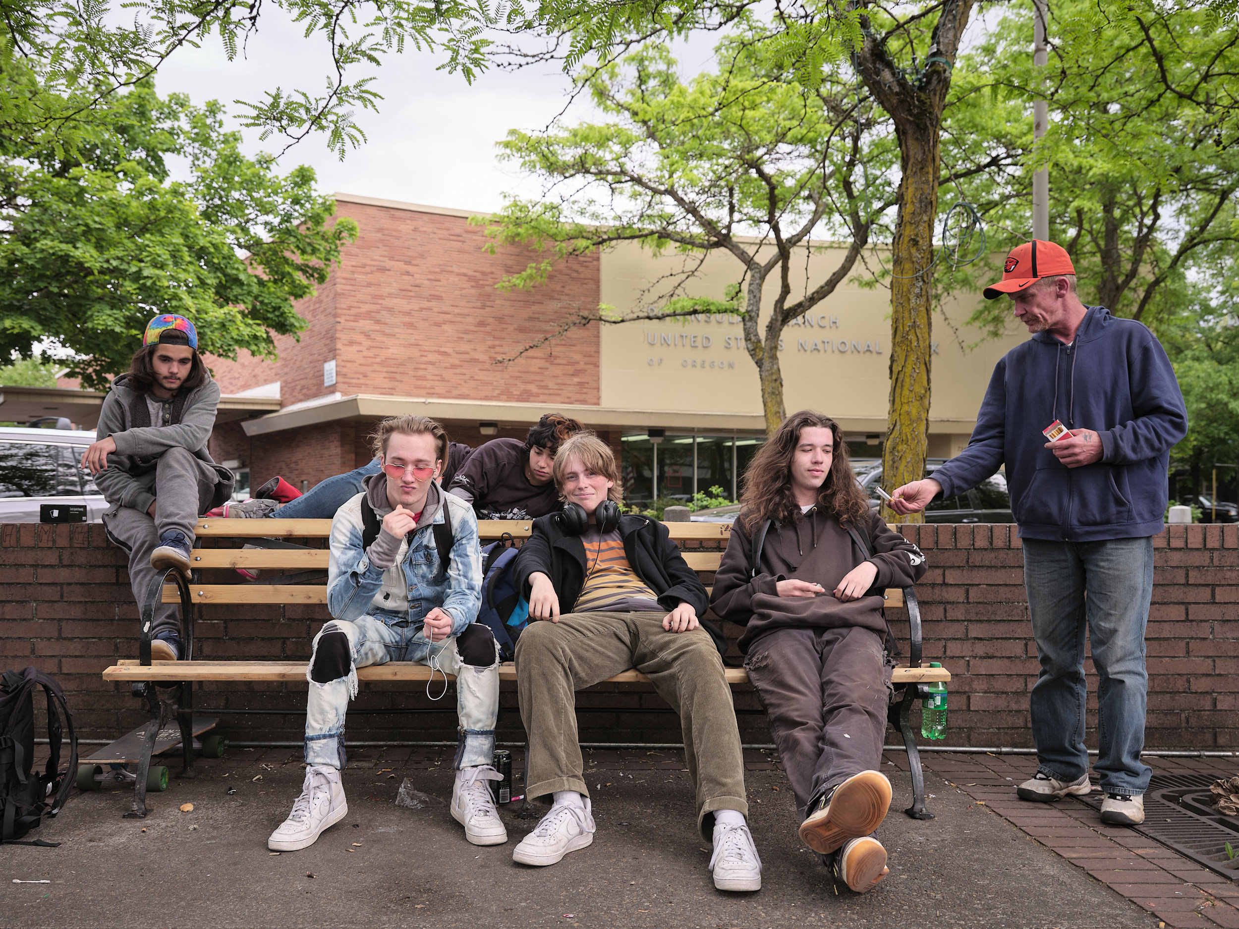 A group of five teenagers hang out on a public bench in the town square of Portland's St. Johns neighborhood. The teens wear a mix of hoodies, ripped jeans, and white sneakers, with skateboards visible beside them. One wears pink-tinted sunglasses, another has headphones around his neck. An older man in an orange Nike cap and blue hoodie stands to the right, offering two cigarettes to one of the teenagers. A brick United States National Bank of Oregon branch building is visible in the background, framed by leafy green trees under an overcast sky.