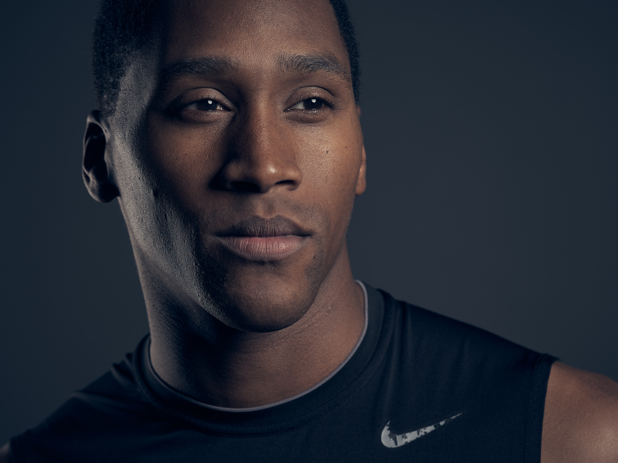 A close-up studio portrait of a young man wearing a black Nike sleeveless shirt, lit dramatically against a dark grey background. He gazes slightly off-camera with a calm, composed expression.