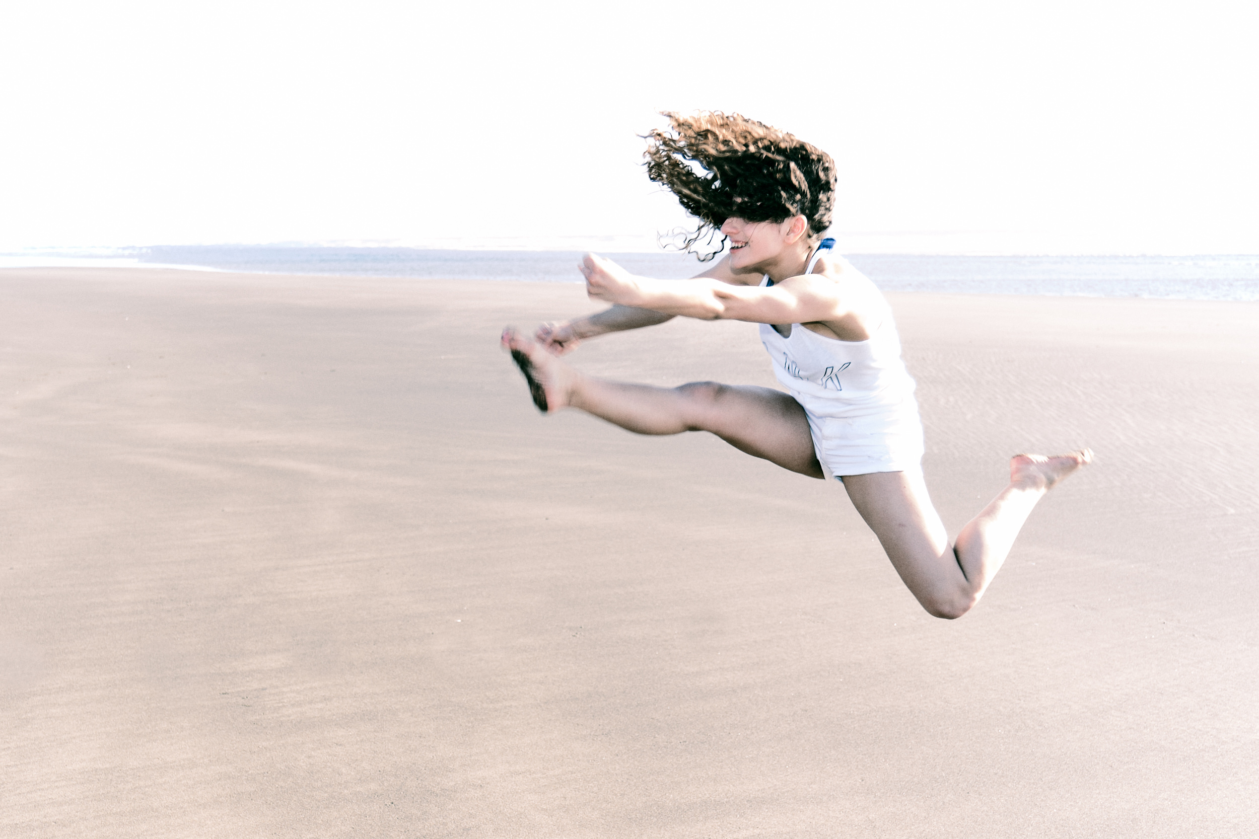 A young woman leaps joyfully into the air on a wide, flat sandy beach, her curly dark hair flying across her face. She wears a white tank top and shorts, arms outstretched and legs bent mid-jump, laughing as she soars above the sand. The bright, overcast sky and pale beach stretch to the horizon behind her.